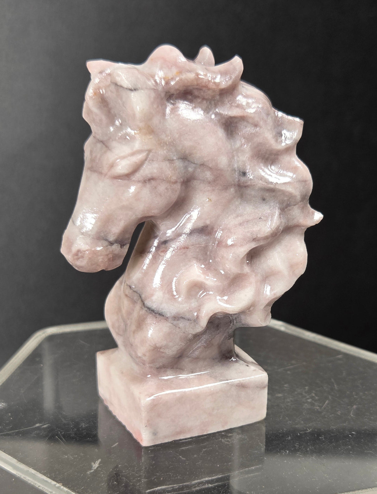 A small sculpted horse head bust made of pale pinkish-gray stone, displayed on a clear acrylic pedestal against a dark background.