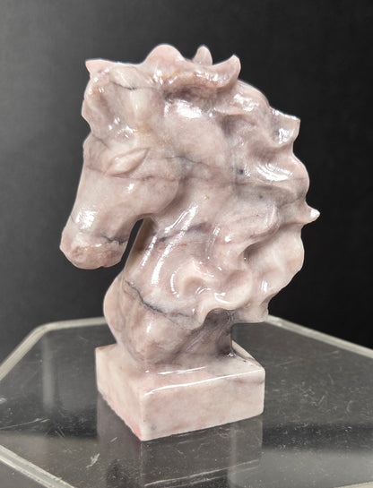A small sculpted horse head bust made of pale pinkish-gray stone, displayed on a clear acrylic pedestal against a dark background.