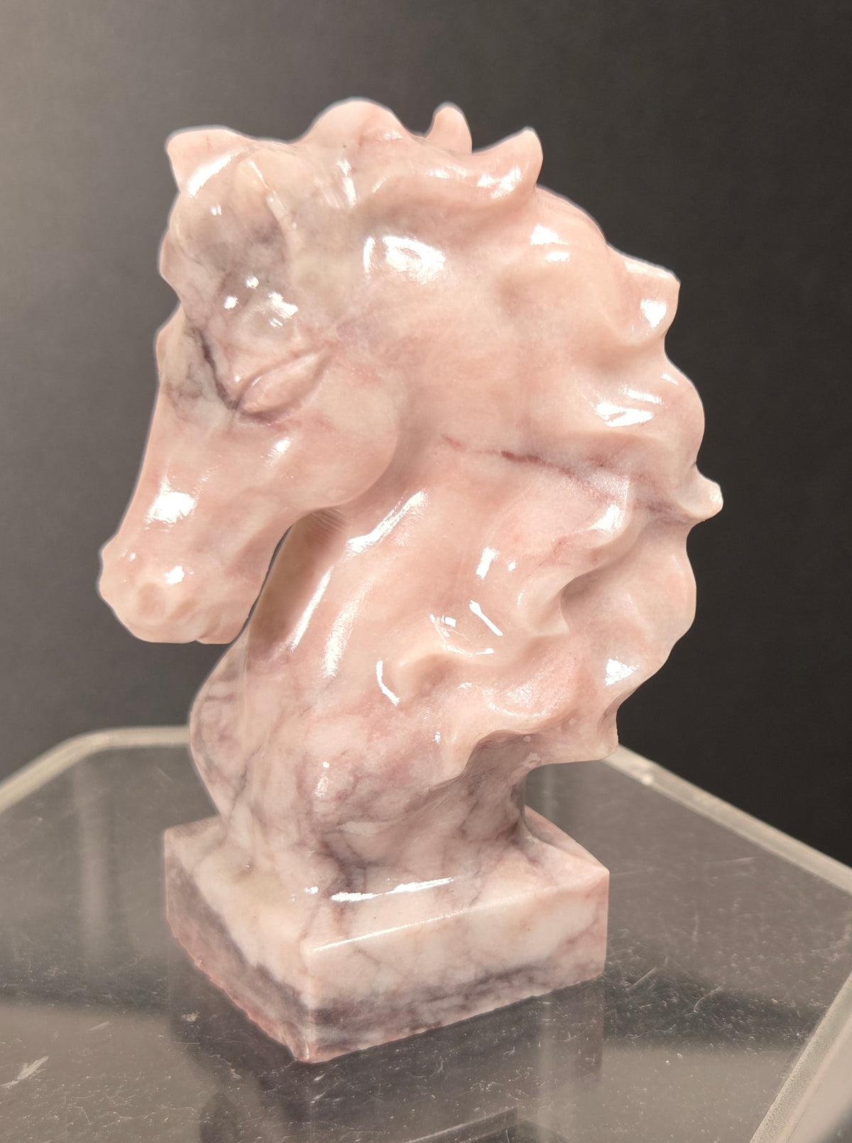 A small polished pink stone horse head sculpture displayed on a clear acrylic stand against a dark gray background.