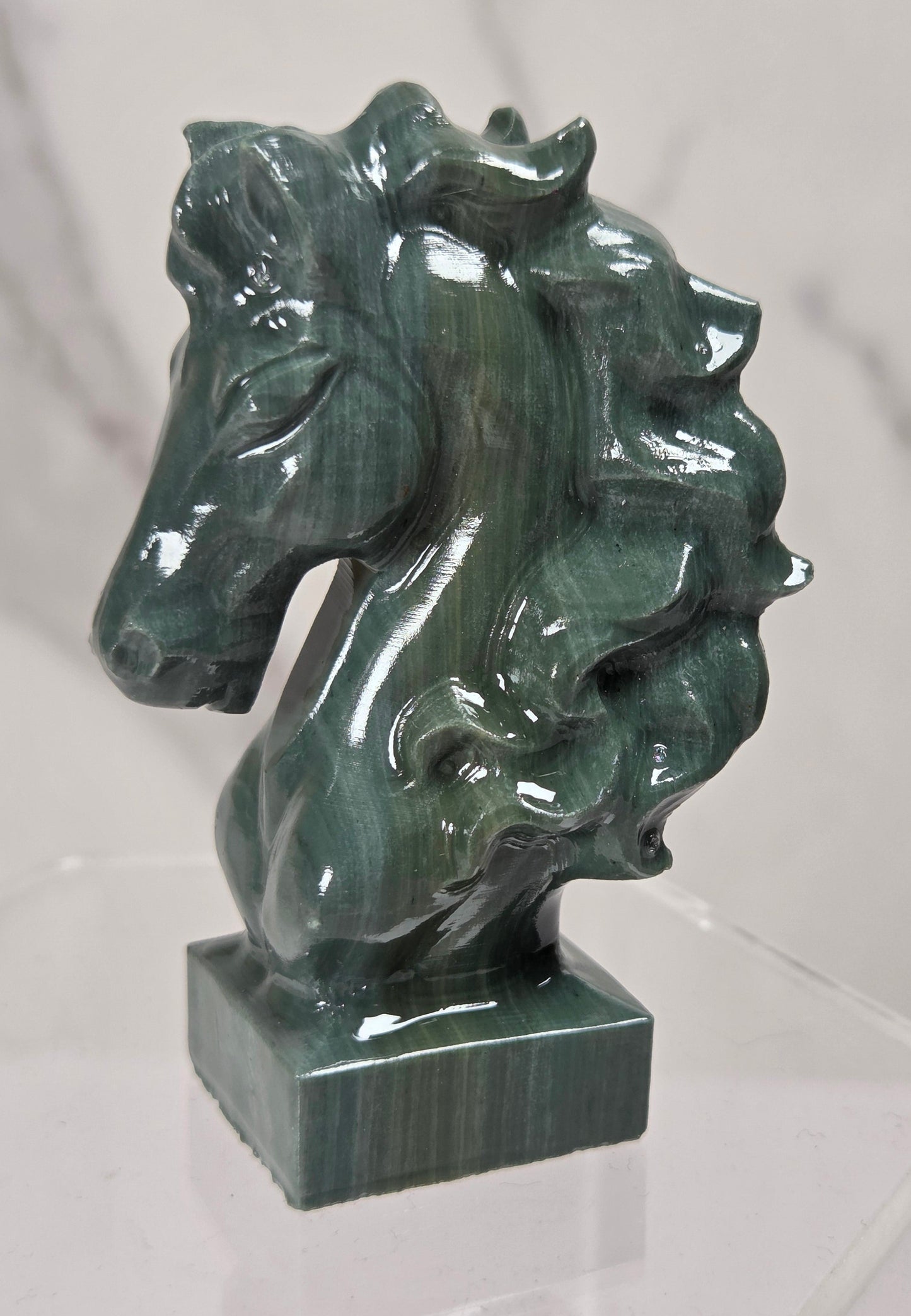 A polished green malachite horse head sculpture on a square base, displayed on a clear acrylic stand against a light gray marble-patterned background.