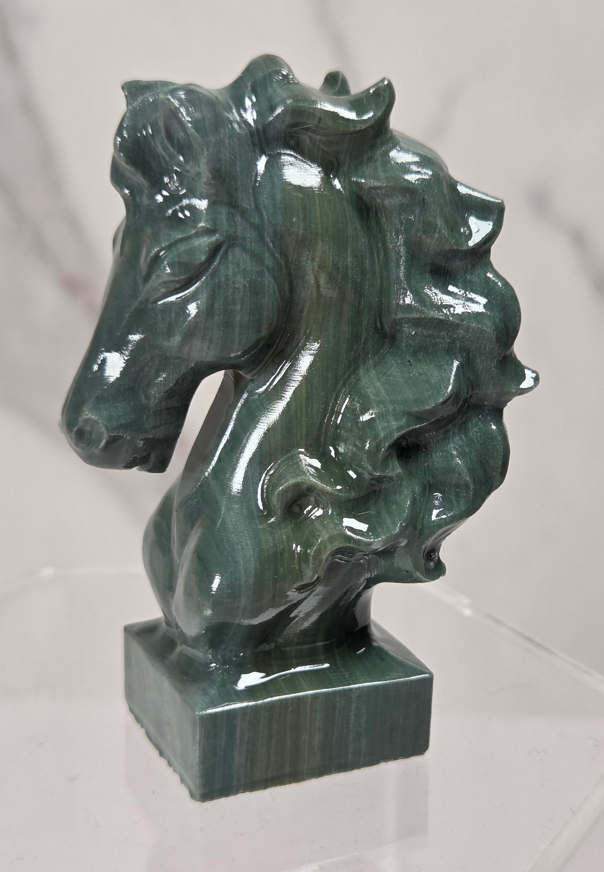 A polished green malachite horse head sculpture on a square base, displayed on a clear acrylic stand against a light gray marble-patterned background.