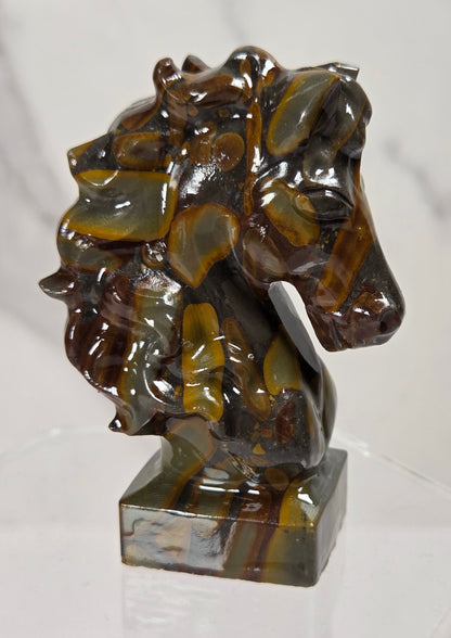 A small sculpted horse head bust made from polished tiger's eye stone, displayed on a clear acrylic stand against a light marble-patterned background.