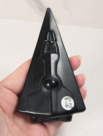 A hand holding a black triangular decorative object with a stylized figure or mask design on the front, photographed against a light indoor background.