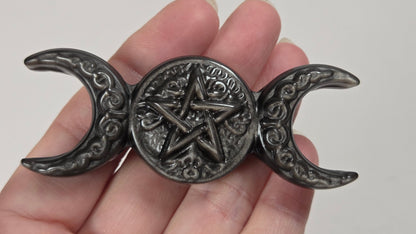 A dark metallic triple moon pentacle figurine held in a hand, featuring an ornate central pentagram within a circular medallion and crescent moon ends with scrollwork details.
