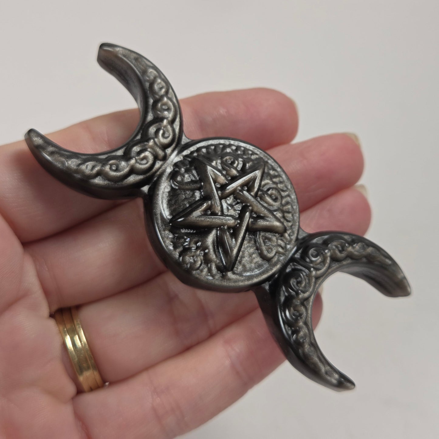 A dark metallic triple moon pentacle figurine held in a hand, featuring an ornate central pentagram within a circular medallion and crescent moon ends with scrollwork details.