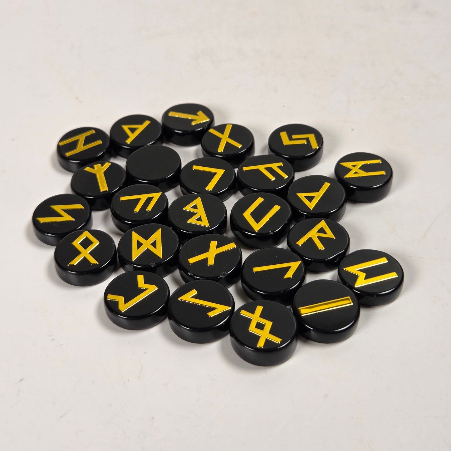 A pile of round black game pawns or tokens printed with yellow runic symbols, arranged on a light off-white surface.