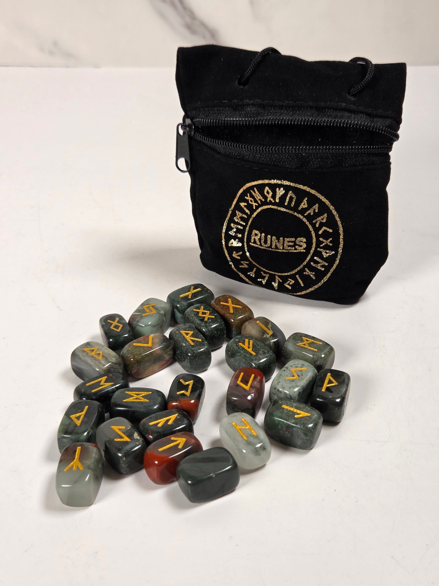 A set of polished tumbled runes stones in various earthy colors, arranged on a white surface in front of a black zippered pouch with a gold 'RUNES' logo.