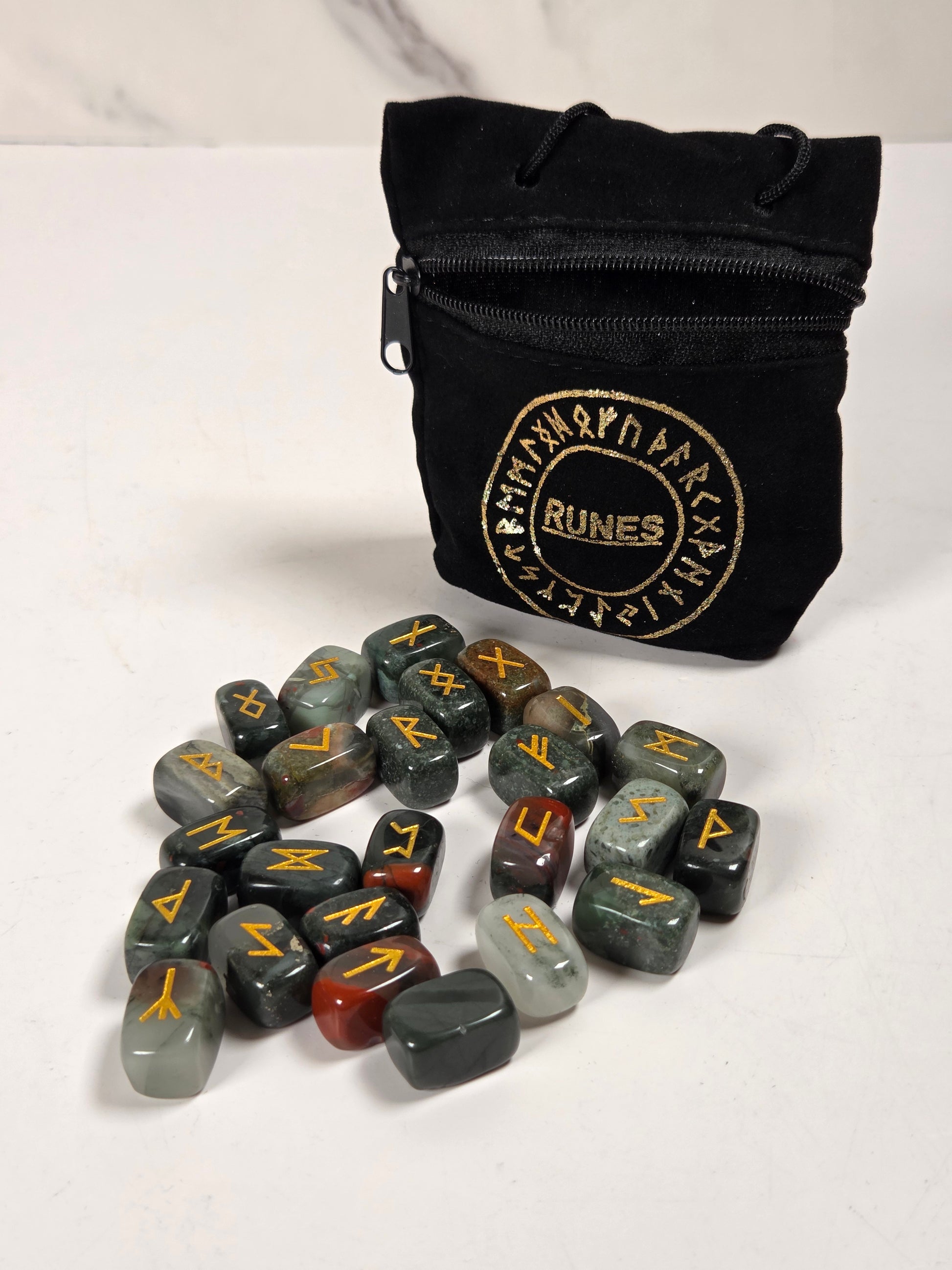 A set of polished tumbled runes stones in various earthy colors, arranged on a white surface in front of a black zippered pouch with a gold 'RUNES' logo.