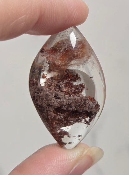 A hand holding a teardrop-shaped clear crystal gemstone with dark brown and reddish-brown inclusions, set against a soft, light gray marble background.