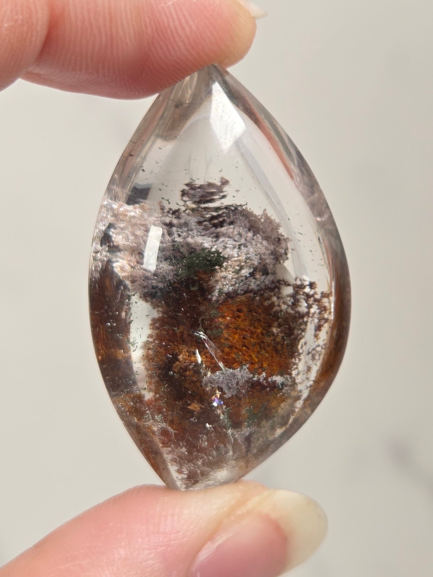 A hand holding a teardrop-shaped clear gemstone with dark brown and black inclusions inside, set against a light gray marble background.