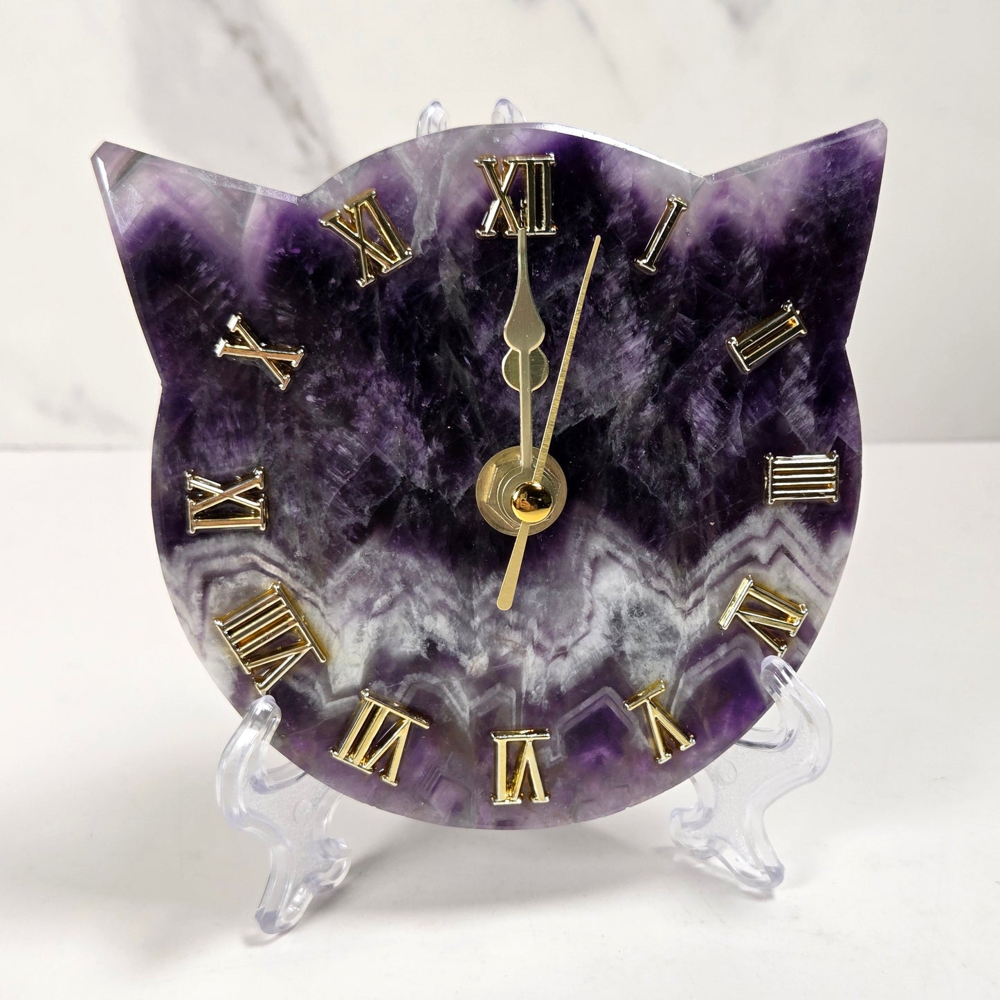 A cat-shaped clock carved from purple and white amethyst with gold Roman numerals and gold hands, displayed on a clear acrylic stand against a white marble background.
