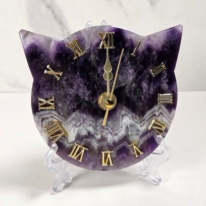 A cat-shaped clock carved from purple and white amethyst with gold Roman numerals and gold hands, displayed on a clear acrylic stand against a white marble background.