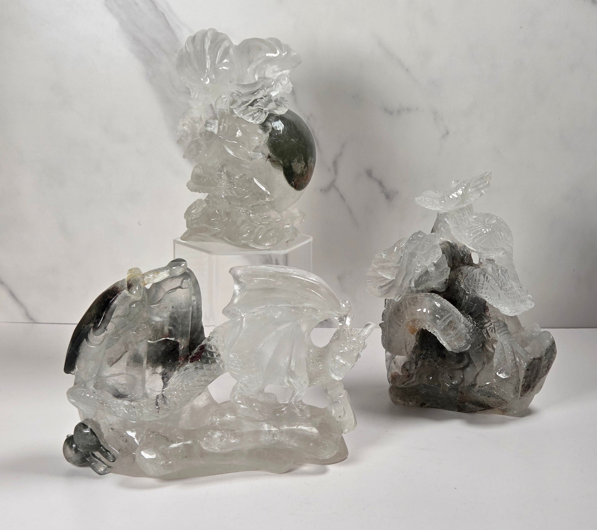 Three carved crystal and stone figurines displayed on a white surface against a white marble-patterned background. The set includes a tall translucent bird figurine on a clear stand, a larger fish figurine with dark inclusions, and a smaller gray stone animal figurine.