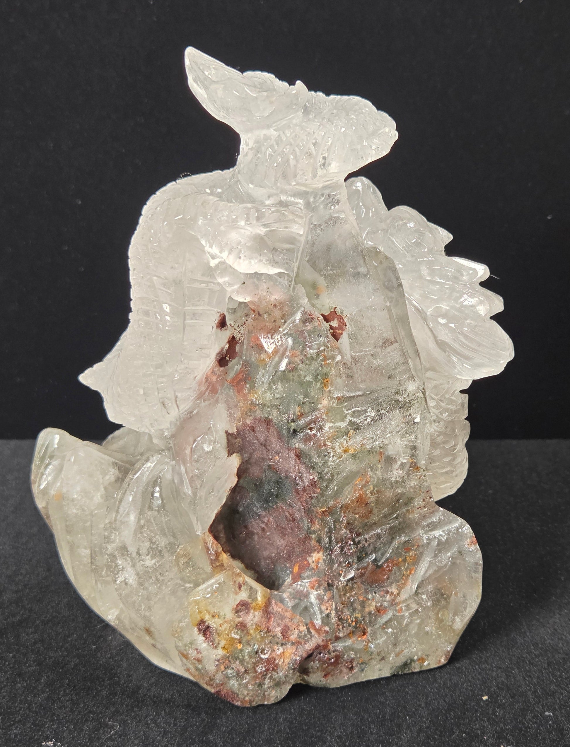 A carved crystal figurine depicting a seated figure, made from transparent clear quartz with natural inclusions and mineral deposits in pink, brown, and green, set against a dark background.