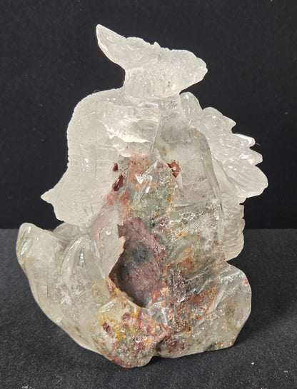 A carved crystal figurine depicting a seated figure, made from transparent clear quartz with natural inclusions and mineral deposits in pink, brown, and green, set against a dark background.
