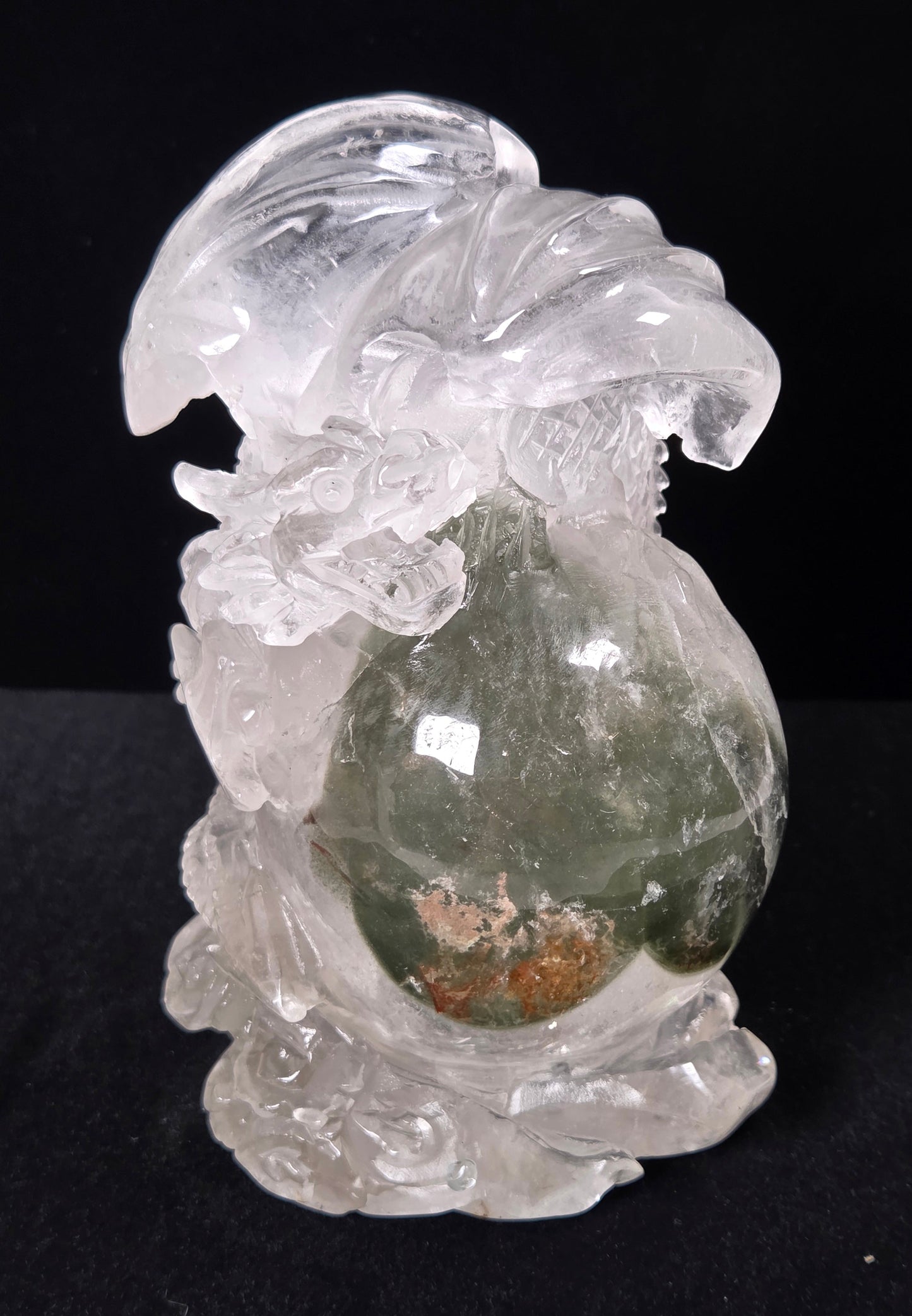 A carved crystal figurine of a dragon holding a green jade-like pouch with a small brown gem inside, set against a solid black background.