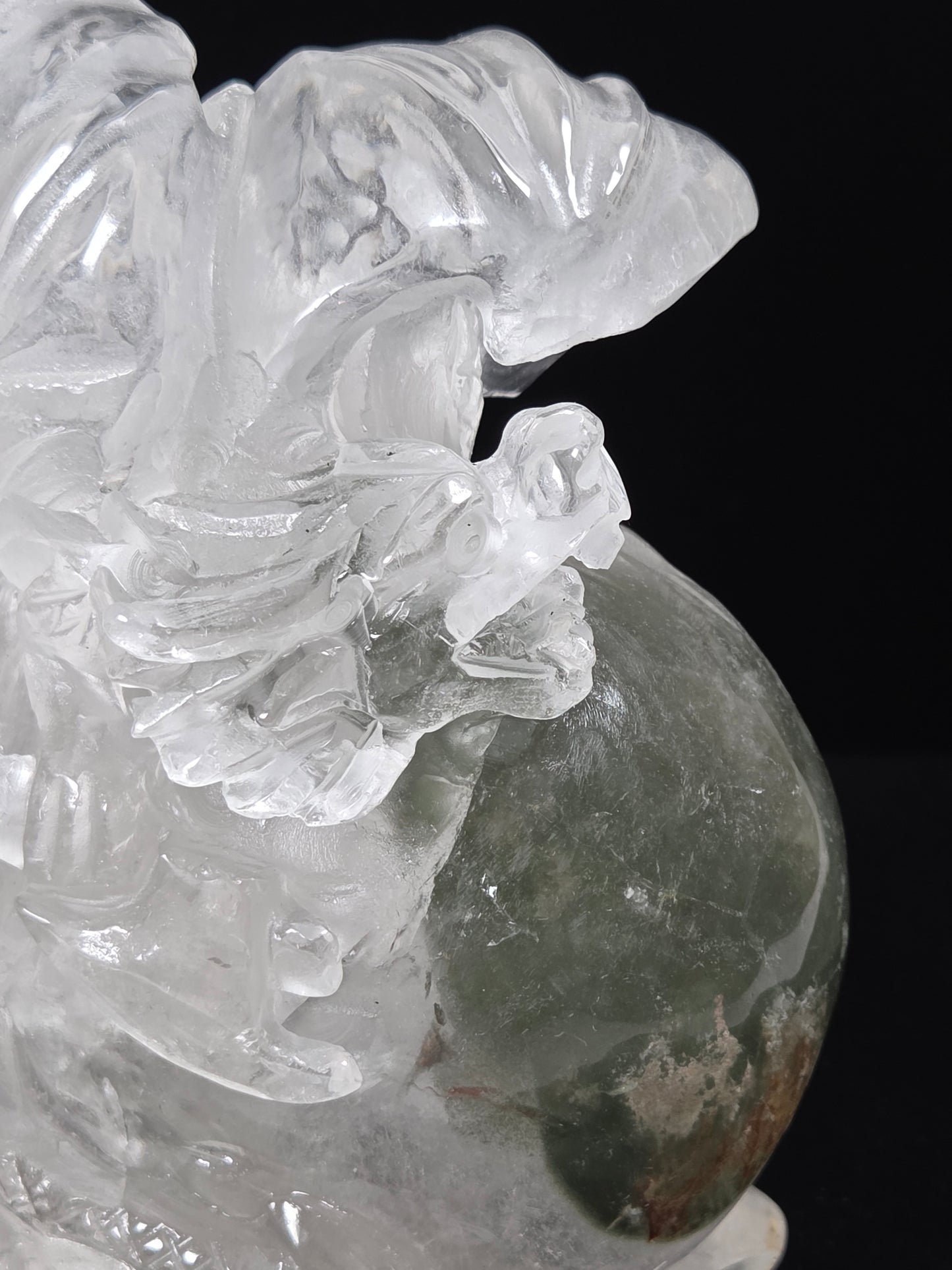 A close-up of a carved crystal figurine depicting a winged deity or animal in transparent clear stone, with a dark green stone sphere at the base, set against a solid black background.