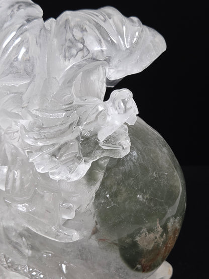 A close-up of a carved crystal figurine depicting a winged deity or animal in transparent clear stone, with a dark green stone sphere at the base, set against a solid black background.