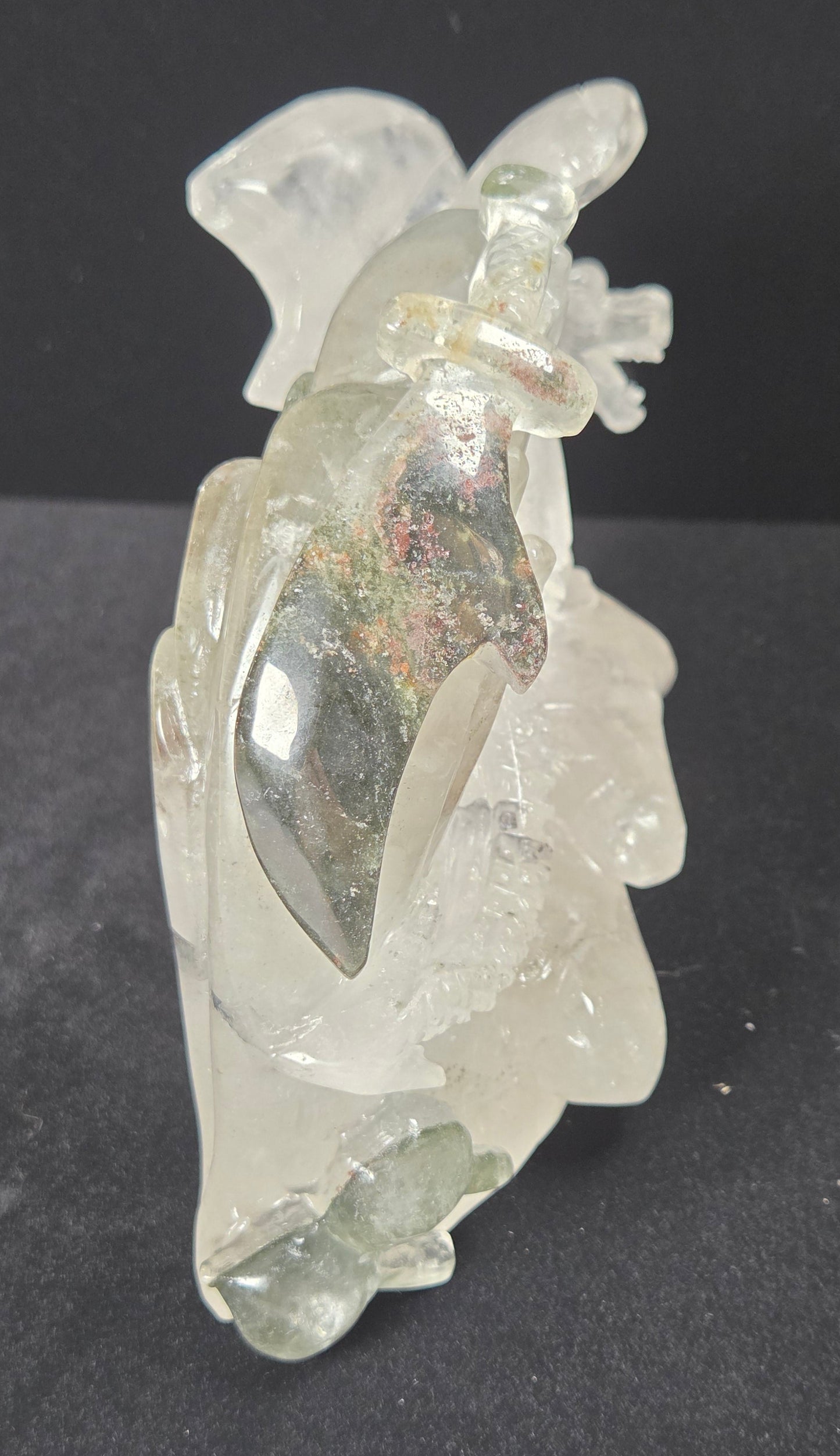 A carved clear crystal figurine of a seated figure holding a sword, displayed on a dark gray background.