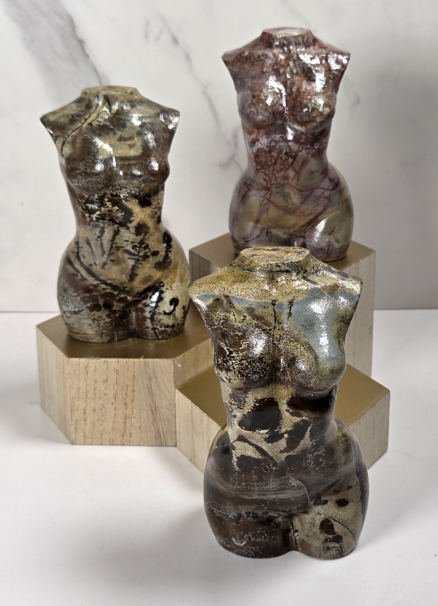 Three small glossy torso sculptures made from marbled stone or resin, each mounted on a hexagonal wooden block, arranged on a white surface against a light marble-look background.
