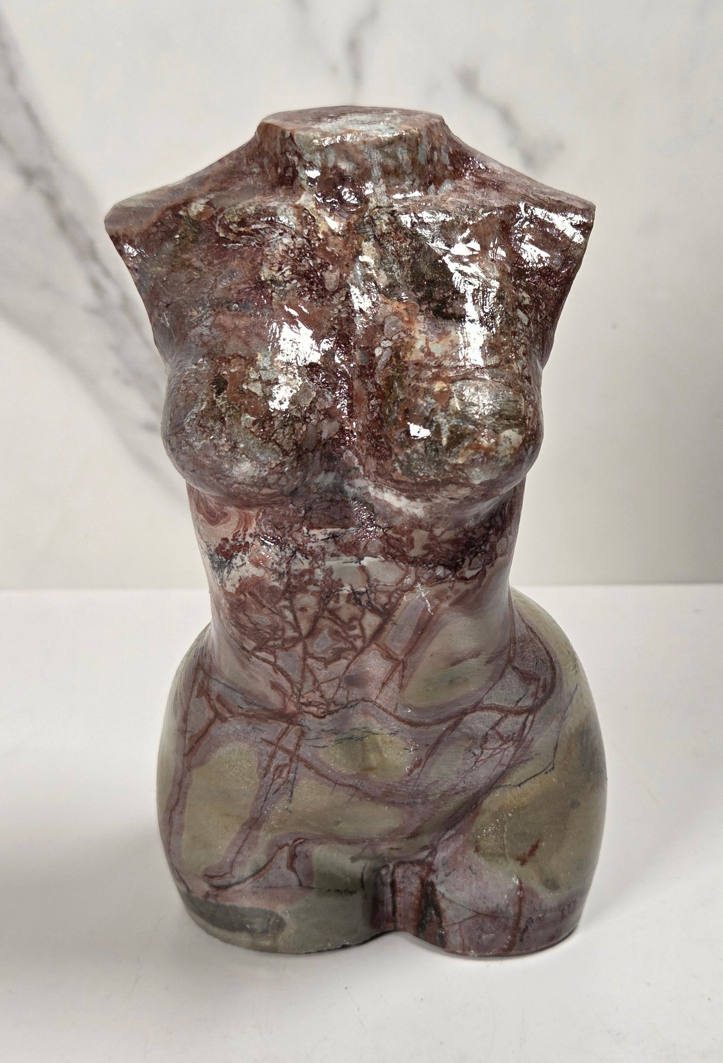 A polished stone torso sculpture with a glossy finish, featuring natural marbled patterns in reddish-brown, gray, and olive green, displayed on a white surface against a light gray marble-patterned background.