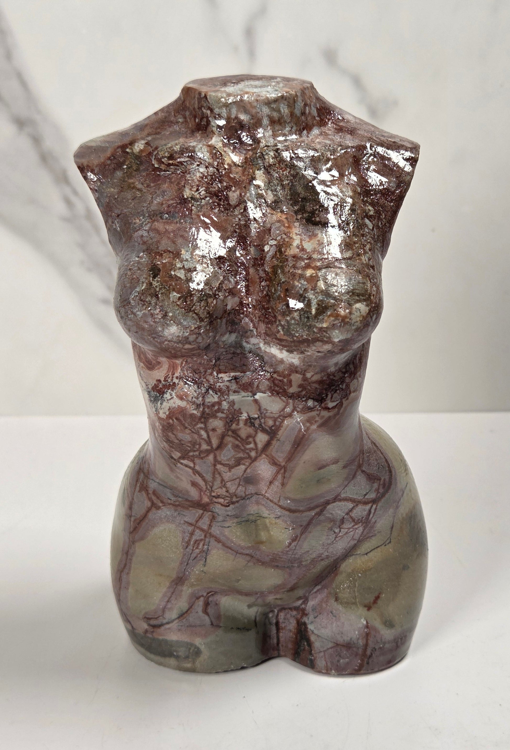 A polished stone torso sculpture with a glossy finish, featuring natural marbled patterns in reddish-brown, gray, and olive green, displayed on a white surface against a light gray marble-patterned background.