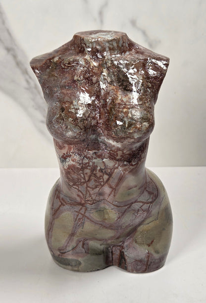 A polished stone torso sculpture with a glossy finish, featuring natural marbled patterns in reddish-brown, gray, and olive green, displayed on a white surface against a light gray marble-patterned background.
