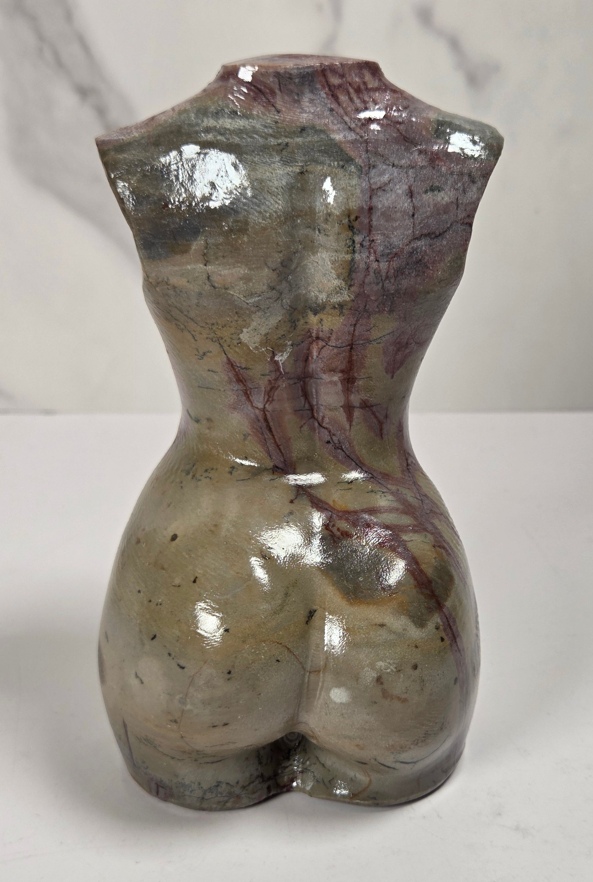 A small sculptural bust of a female torso made from polished stone, showing a glossy marbled surface with gray, beige, and reddish-brown veining, photographed on a white surface against a light marble-look background.