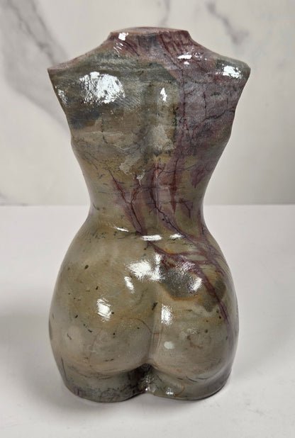 A small sculptural bust of a female torso made from polished stone, showing a glossy marbled surface with gray, beige, and reddish-brown veining, photographed on a white surface against a light marble-look background.