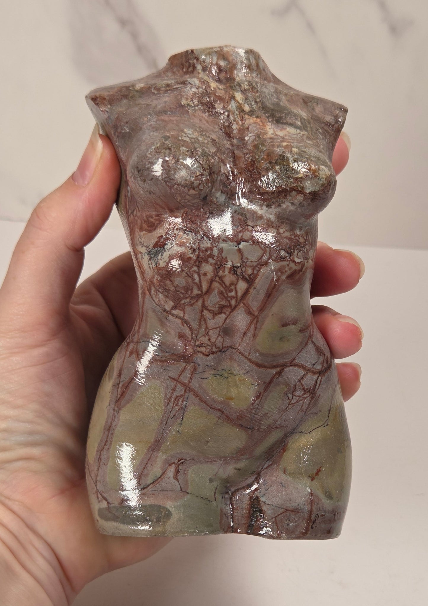 A hand holding a small glossy ceramic torso sculpture with a mottled earthy color palette of brown, gray, and muted green, set against a light gray marble-patterned background.