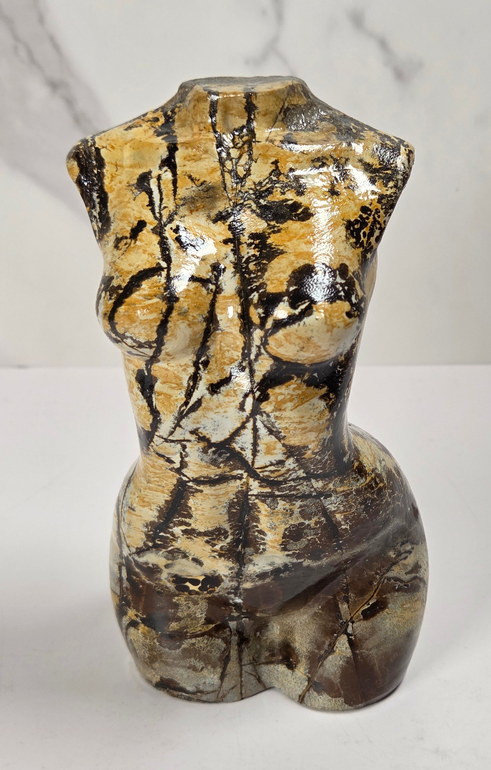 A small sculptural torso-shaped vase with a glossy marbled finish in beige, brown, and black, displayed on a white surface against a light gray marble-patterned background.