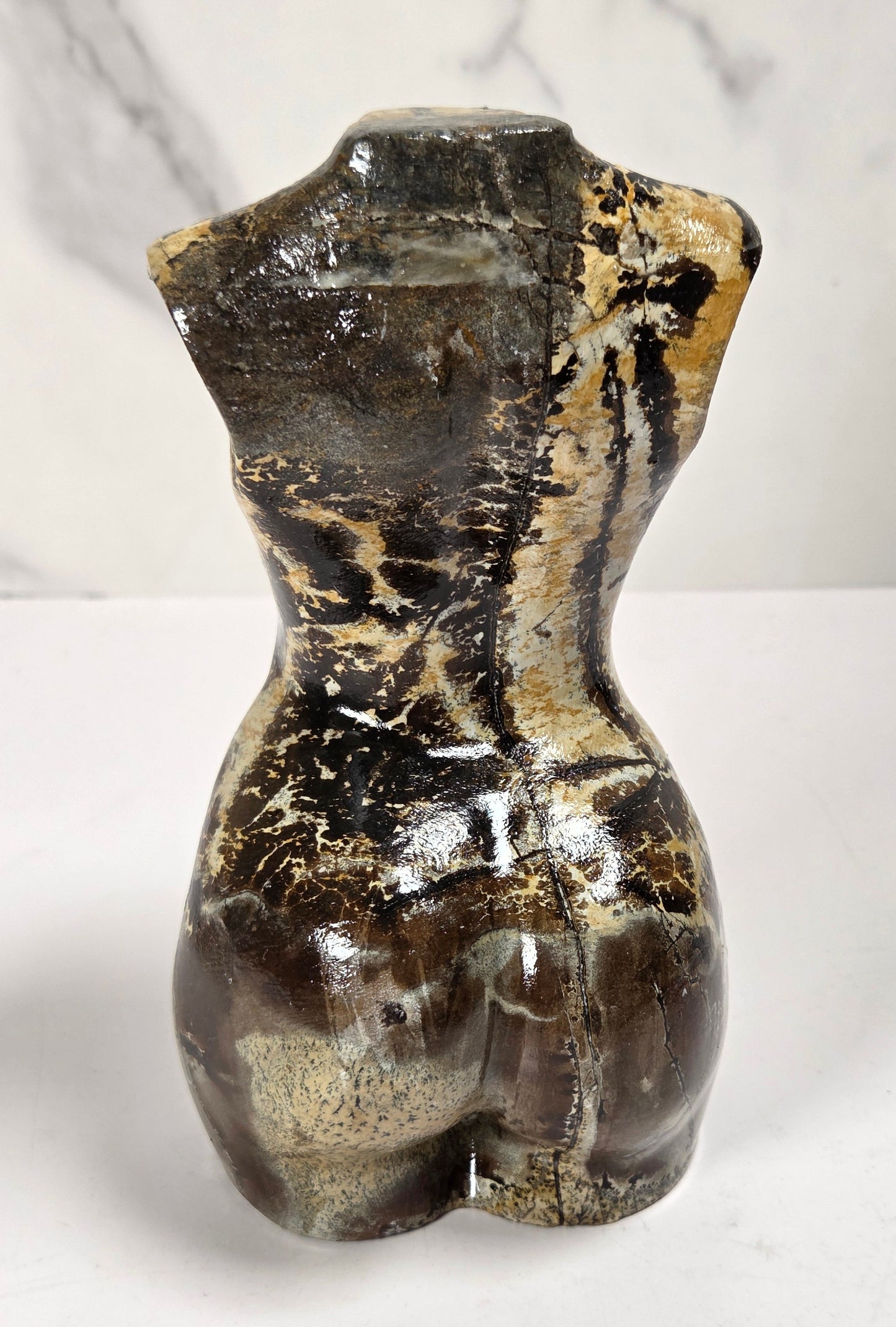 A small sculptural torso form resembling a female waist and hips, made from polished brown and black stone with beige marbling and a glossy finish, standing on a white surface against a white marble-patterned background.