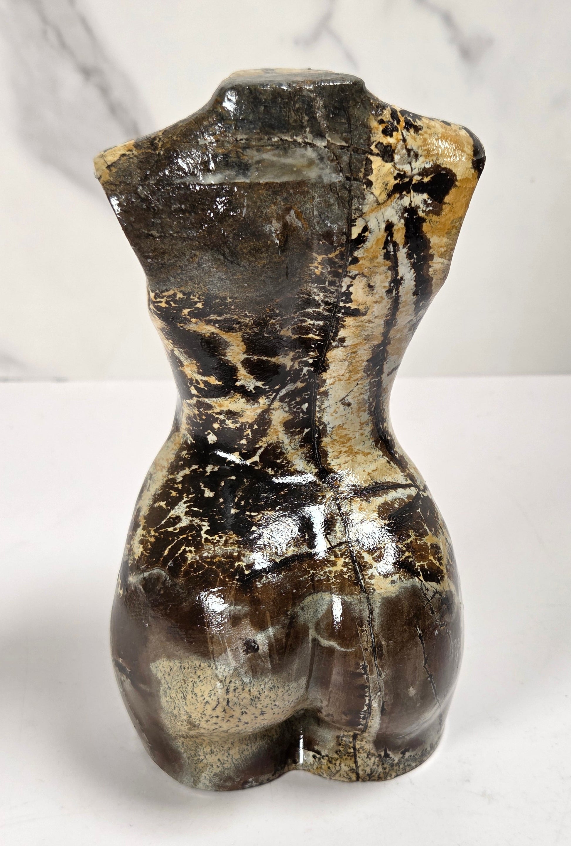 A small sculptural torso form resembling a female waist and hips, made from polished brown and black stone with beige marbling and a glossy finish, standing on a white surface against a white marble-patterned background.