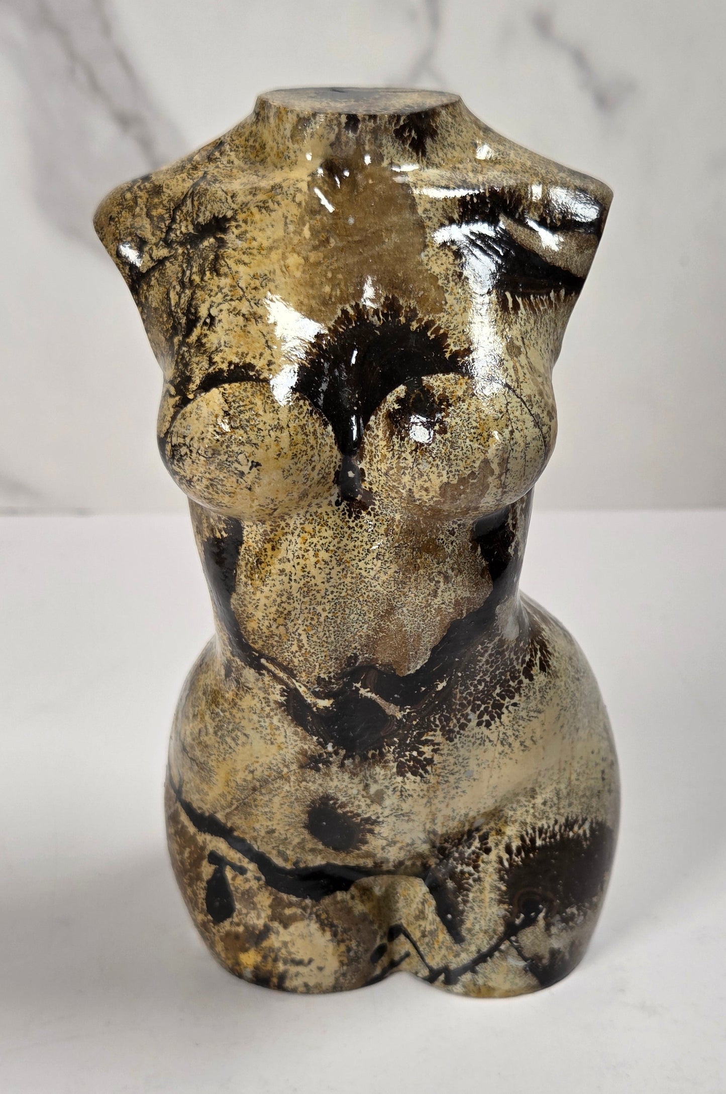 A small sculptural torso-shaped carving with a glossy marbled finish in beige, brown, and black, displayed on a white surface against a light gray marble-patterned background.