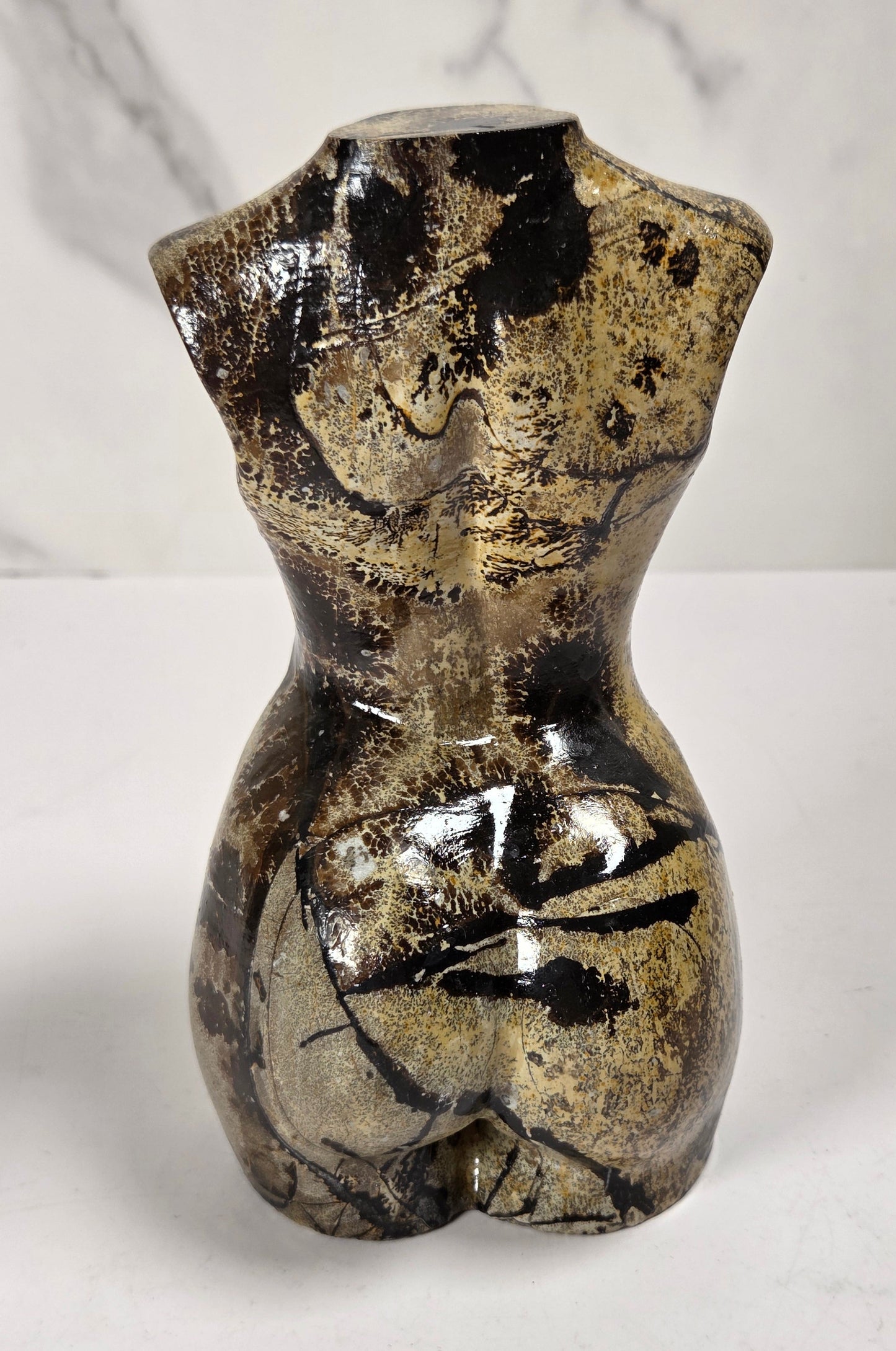 A small sculptural vase shaped like a stylized female torso, with a glossy black and metallic gold marbled pattern, set against a white marble-patterned background.