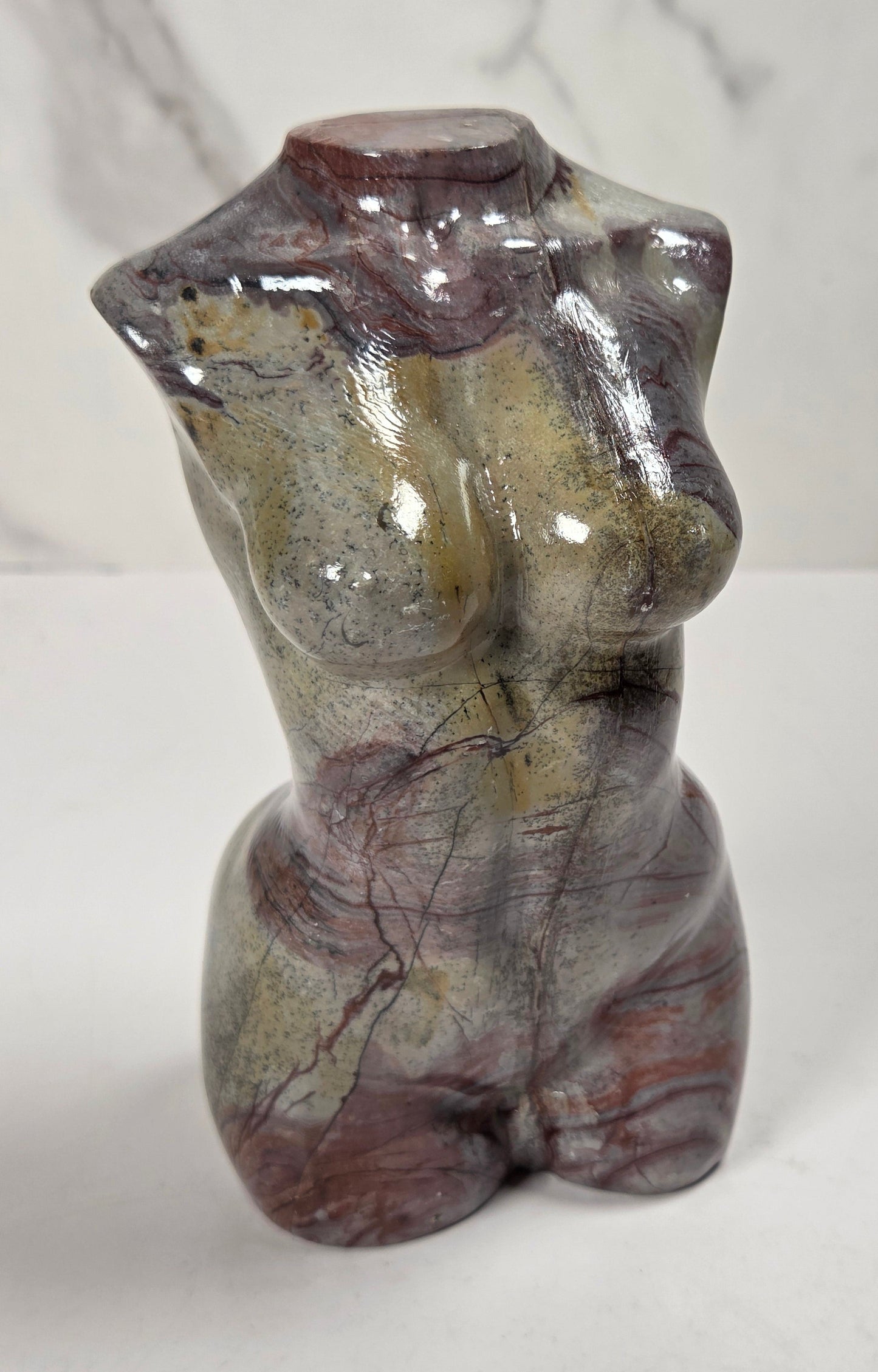 A small glossy stone torso sculpture with a stylized female form, featuring natural marbled patterns in brown, gray, and muted green, set against a light gray marble-patterned background.