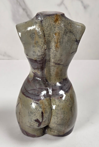A small sculptural female torso bust made of polished stone or ceramic, shown from the back on a white surface with a light gray marble-look background.