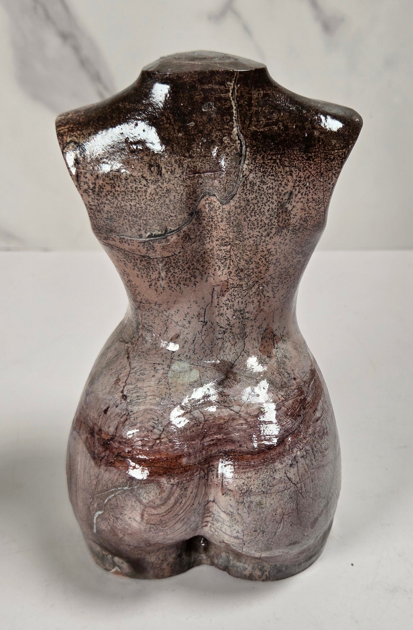 A glossy ceramic female torso sculpture in a mottled brown and gray glaze, shown from the back on a light gray surface with a marble-patterned background.