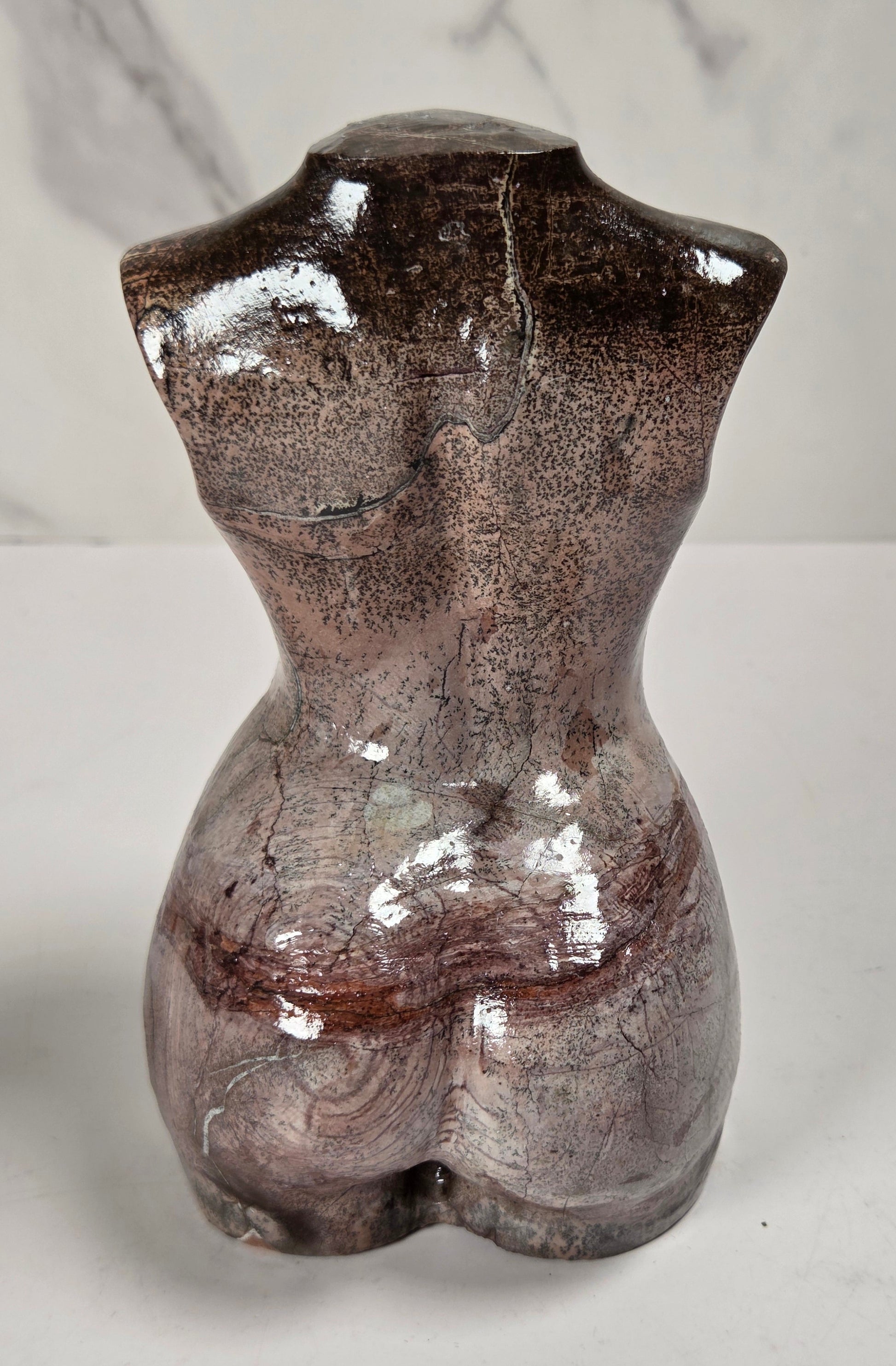 A glossy ceramic female torso sculpture in a mottled brown and gray glaze, shown from the back on a light gray surface with a marble-patterned background.