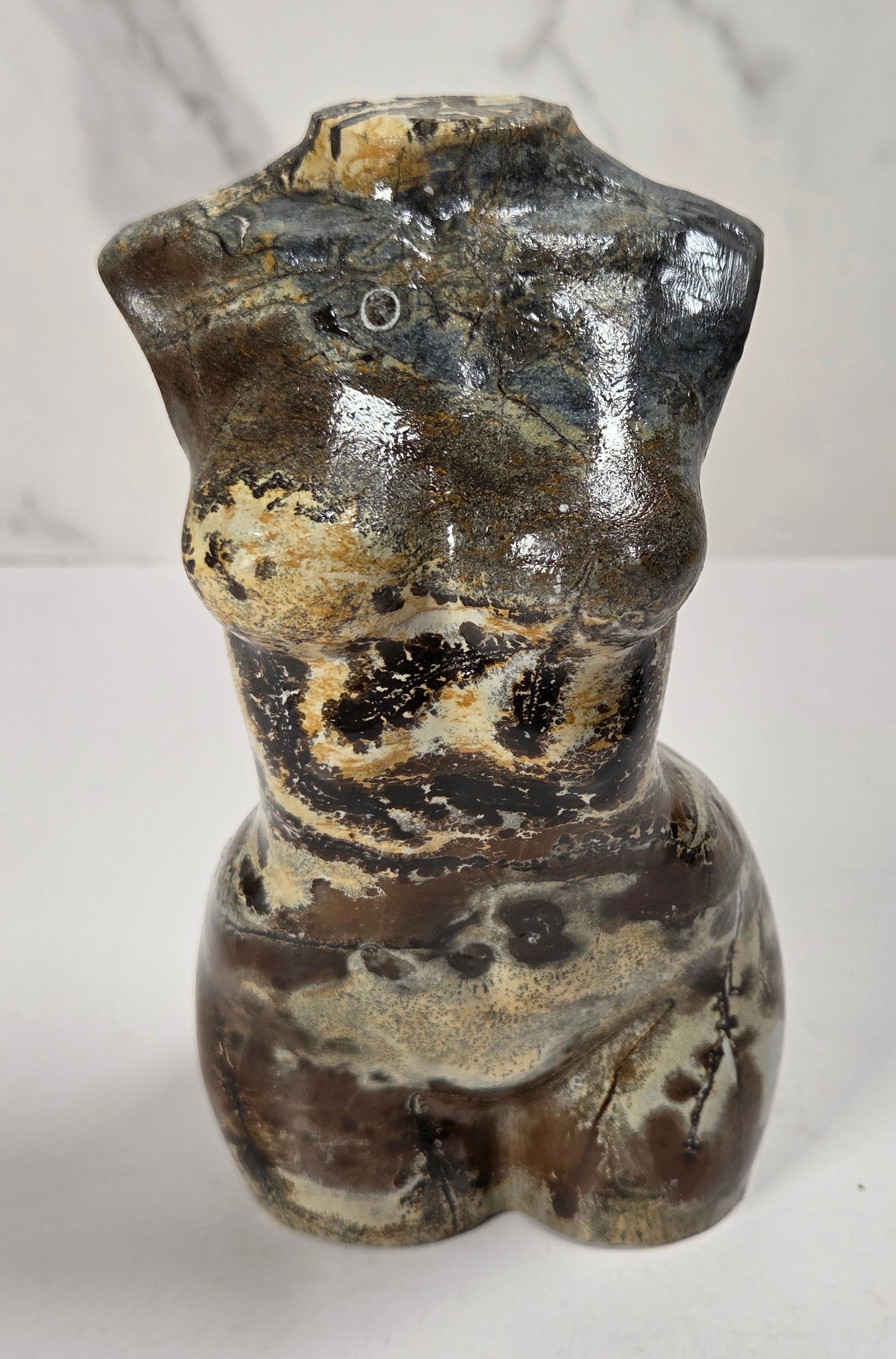 A small sculptural torso figurine with a glossy glaze, featuring mottled brown, black, beige, and gray tones, set against a light gray marble-patterned background.