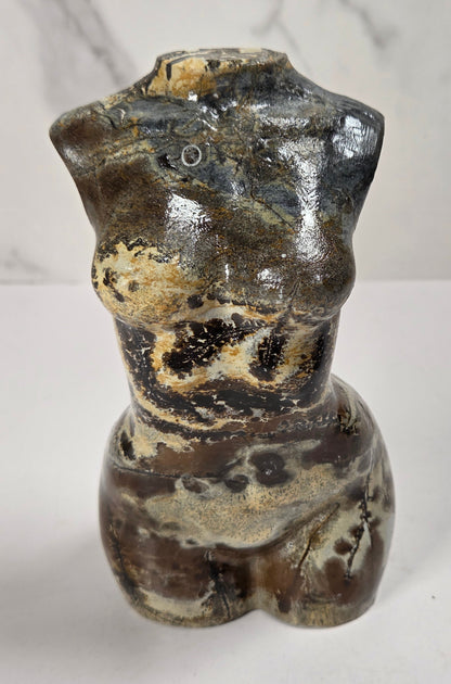 A small sculptural torso figurine with a glossy glaze, featuring mottled brown, black, beige, and gray tones, set against a light gray marble-patterned background.