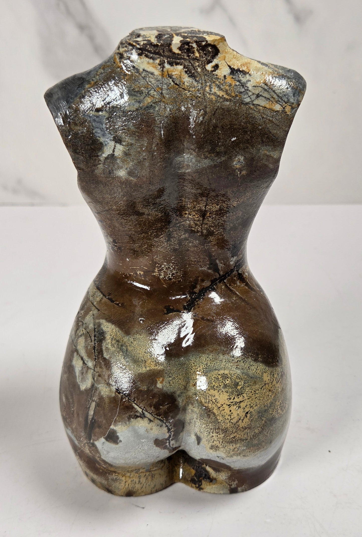 A small glossy ceramic female torso sculpture with a textured, marbled finish in dark brown, black, and metallic gold, photographed against a light gray marble-patterned background.