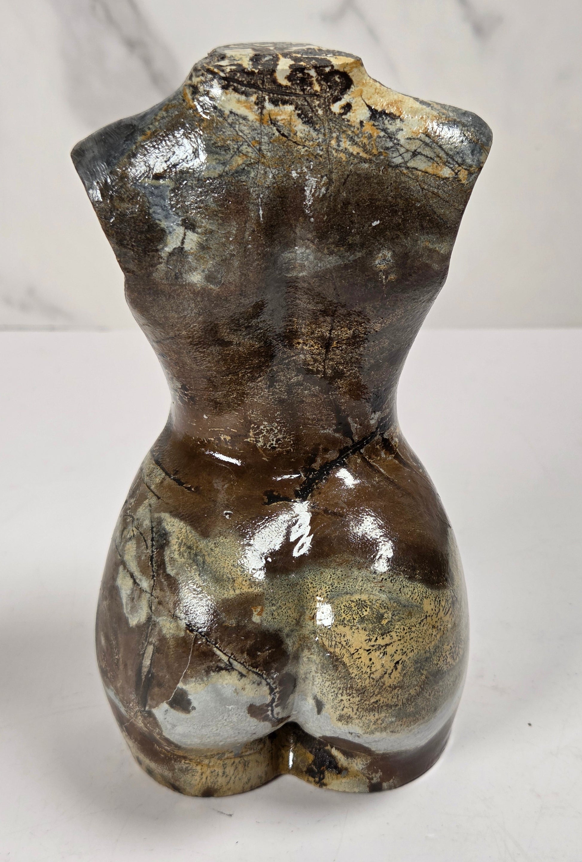 A small glossy ceramic female torso sculpture with a textured, marbled finish in dark brown, black, and metallic gold, photographed against a light gray marble-patterned background.