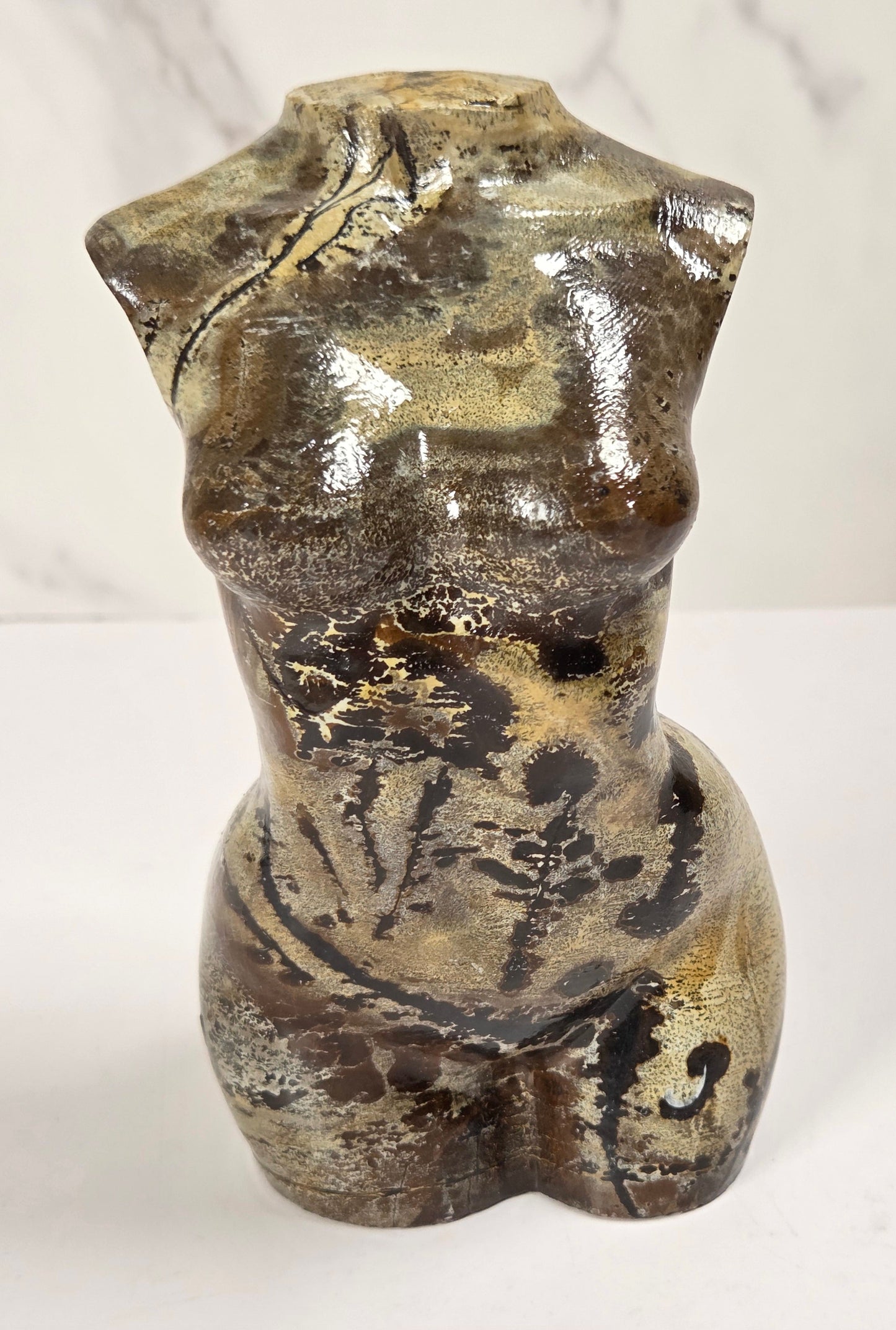 A small sculptural torso-shaped vase or decorative object with a glossy abstract finish in earthy brown, beige, and black tones, photographed against a light marble-patterned background.