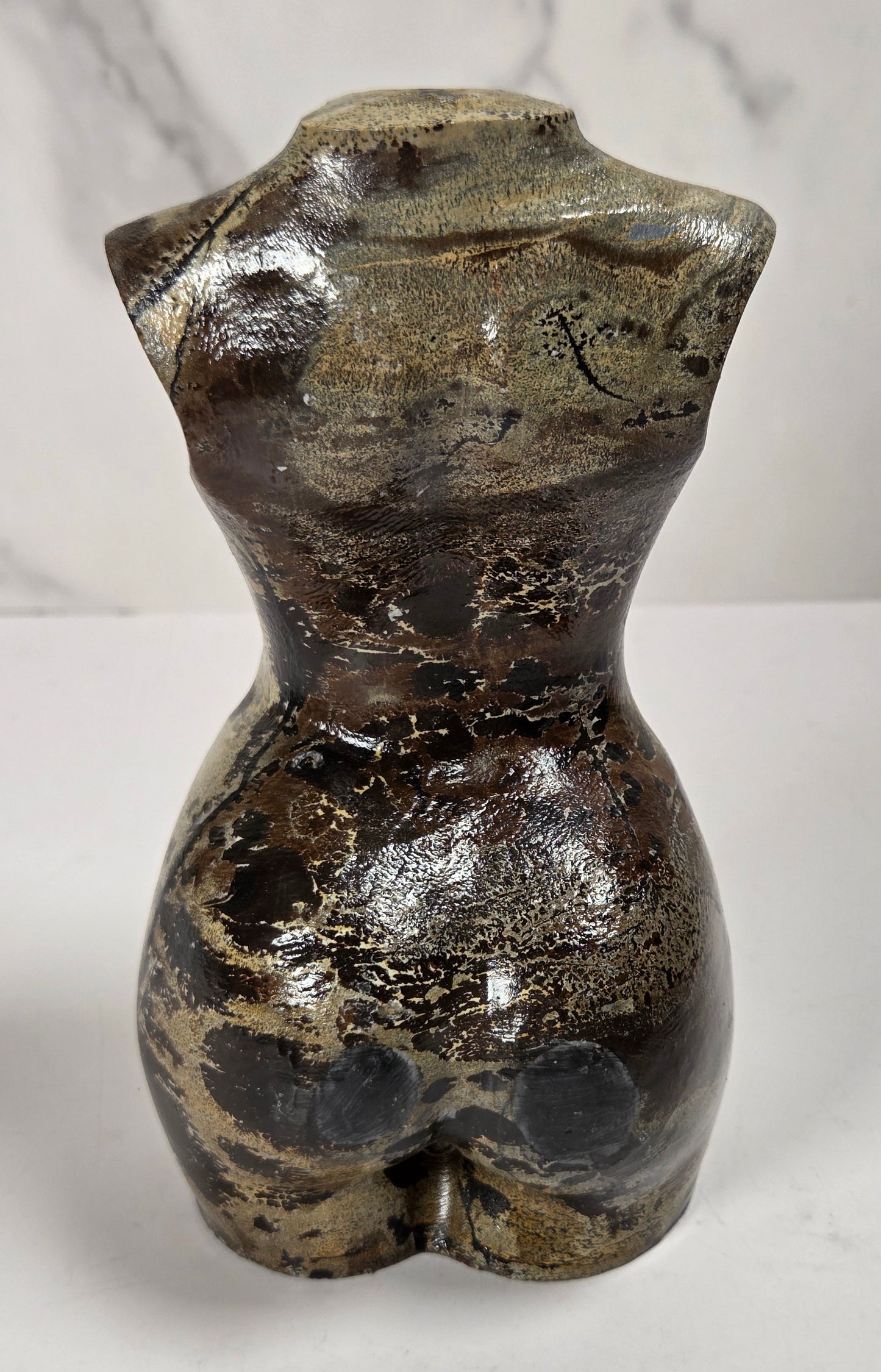 A small sculptural torso-shaped carving with a glossy marbled finish in beige, brown, and black, displayed on a white surface against a light gray marble-patterned background.