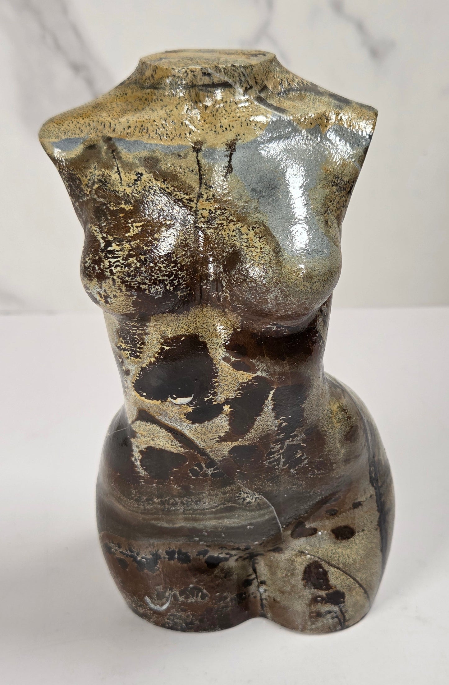 A small sculptural torso-shaped ceramic vase with a glossy glaze in mottled brown, beige, and gray tones, photographed on a white surface against a light marble-patterned background.