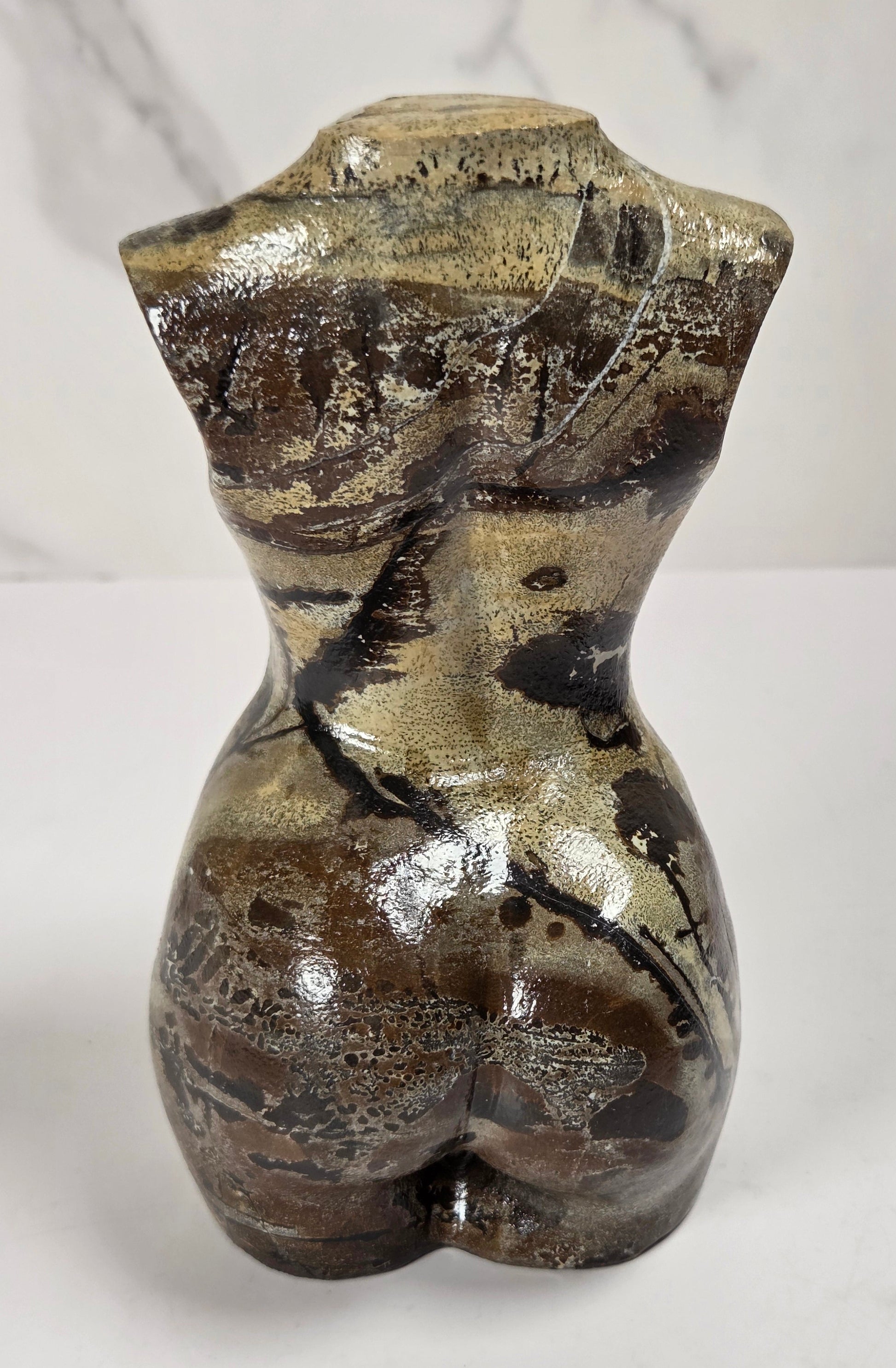 A sculptural female torso figurine with a glossy, metallic finish in shades of bronze, gold, and dark brown, set against a light gray marble-patterned background.