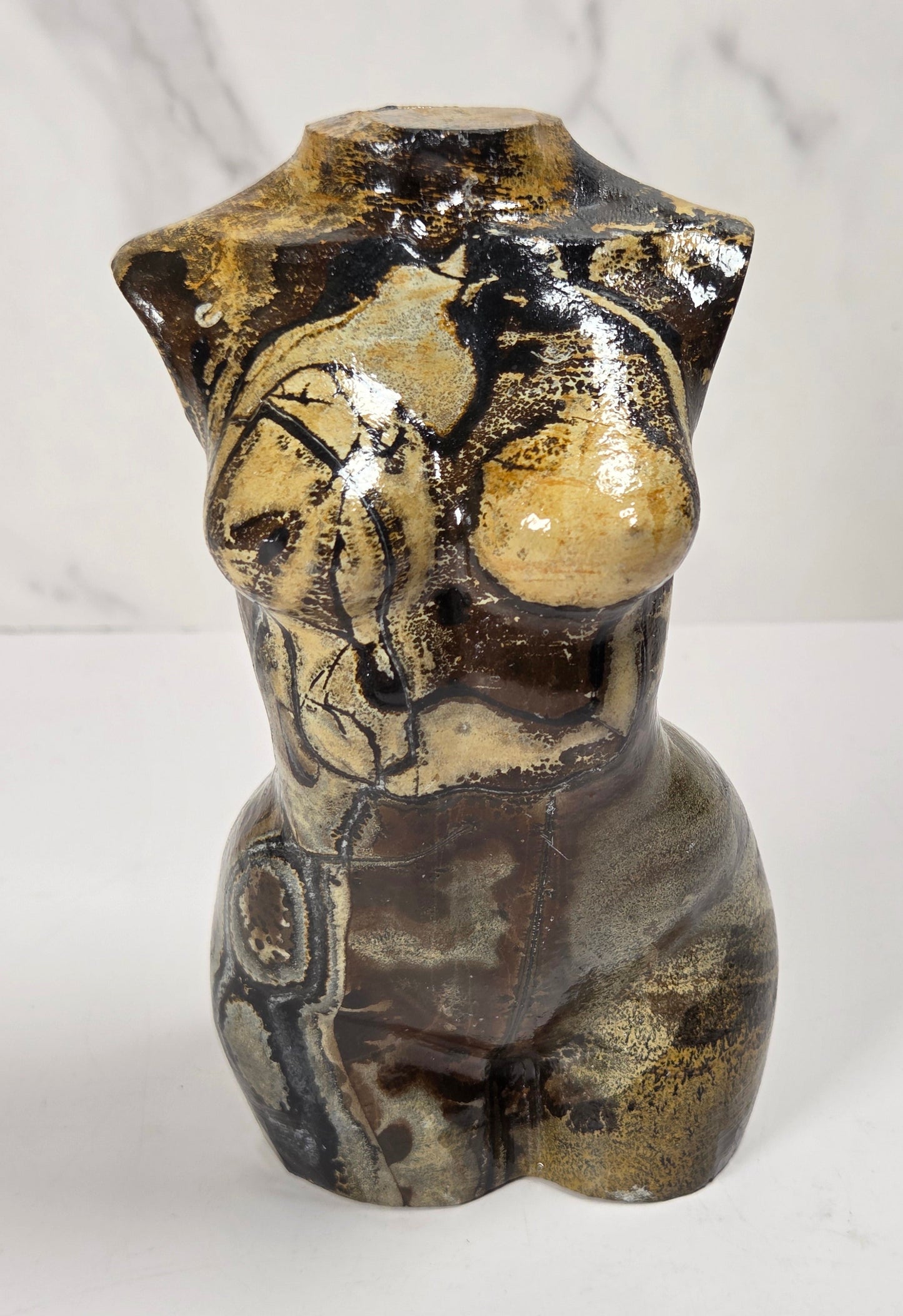 A ceramic decorative sculpture of a stylized female torso with glossy glaze, featuring a mix of black, brown, and beige colors, set against a white marble-patterned background.