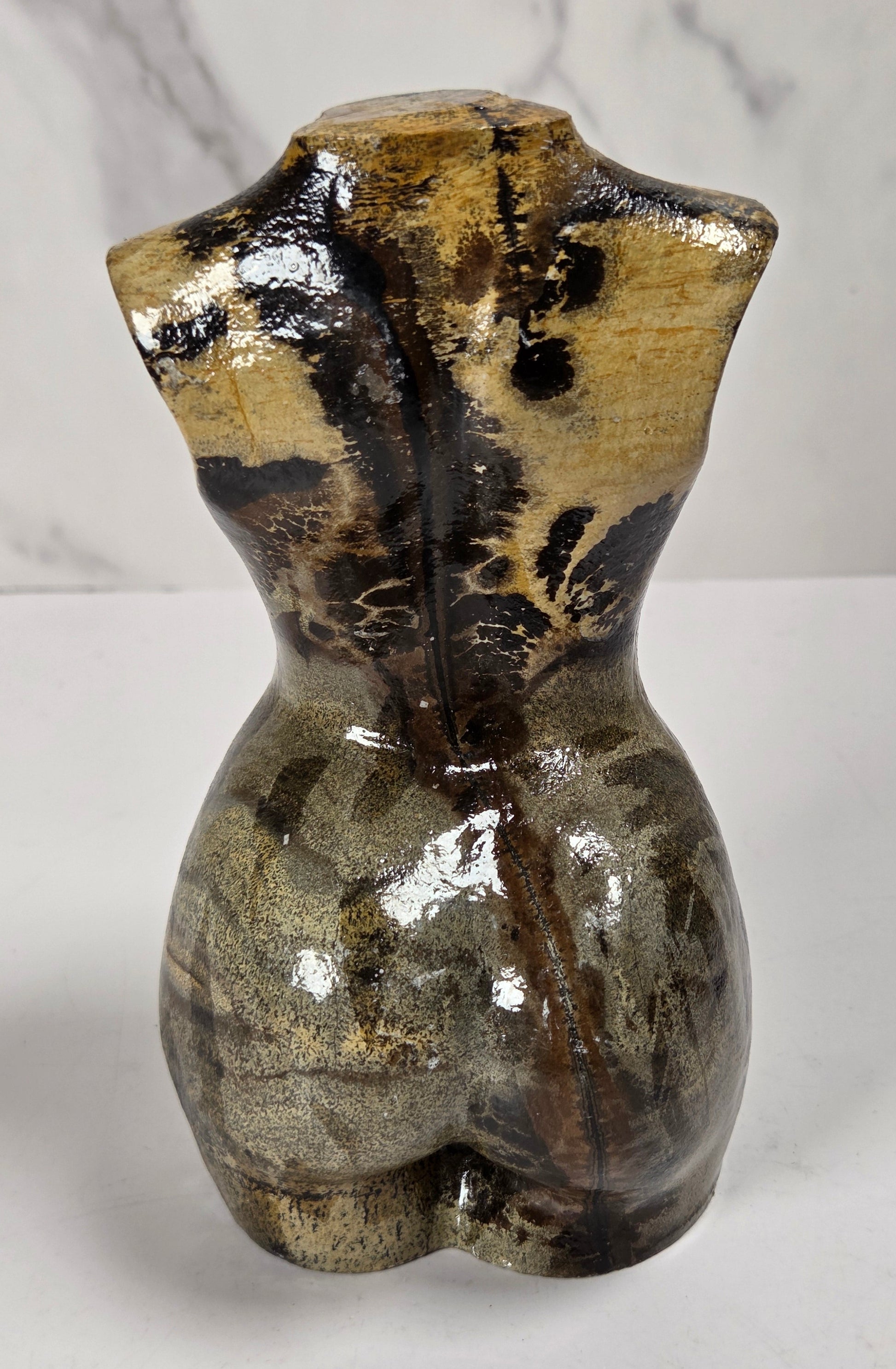 A small sculptural torso-shaped object with a glossy marbled finish in earthy brown, beige, and black tones, photographed against a light gray marble-patterned background.