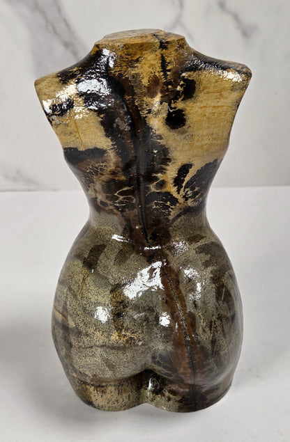 A small sculptural torso-shaped object with a glossy marbled finish in earthy brown, beige, and black tones, photographed against a light gray marble-patterned background.